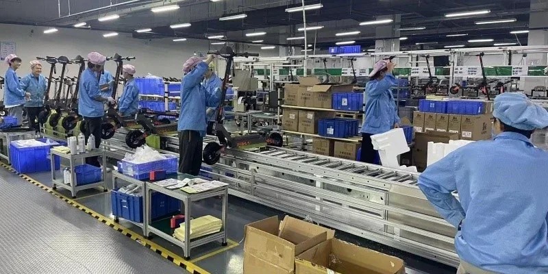 Linia montażowa hulajnóg KuKirin: pracownicy w niebieskich fartuchach przy przenośniku rolkowym, pojemniki z częściami